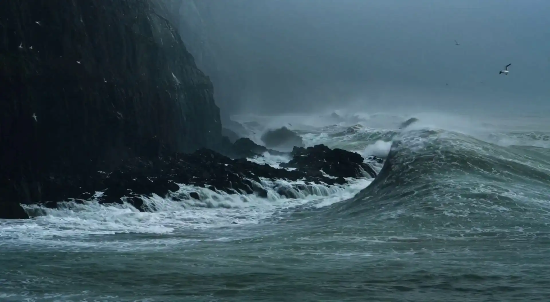 Storm waves breaking against a dark volcanic coastline beneath a blue-gray sky